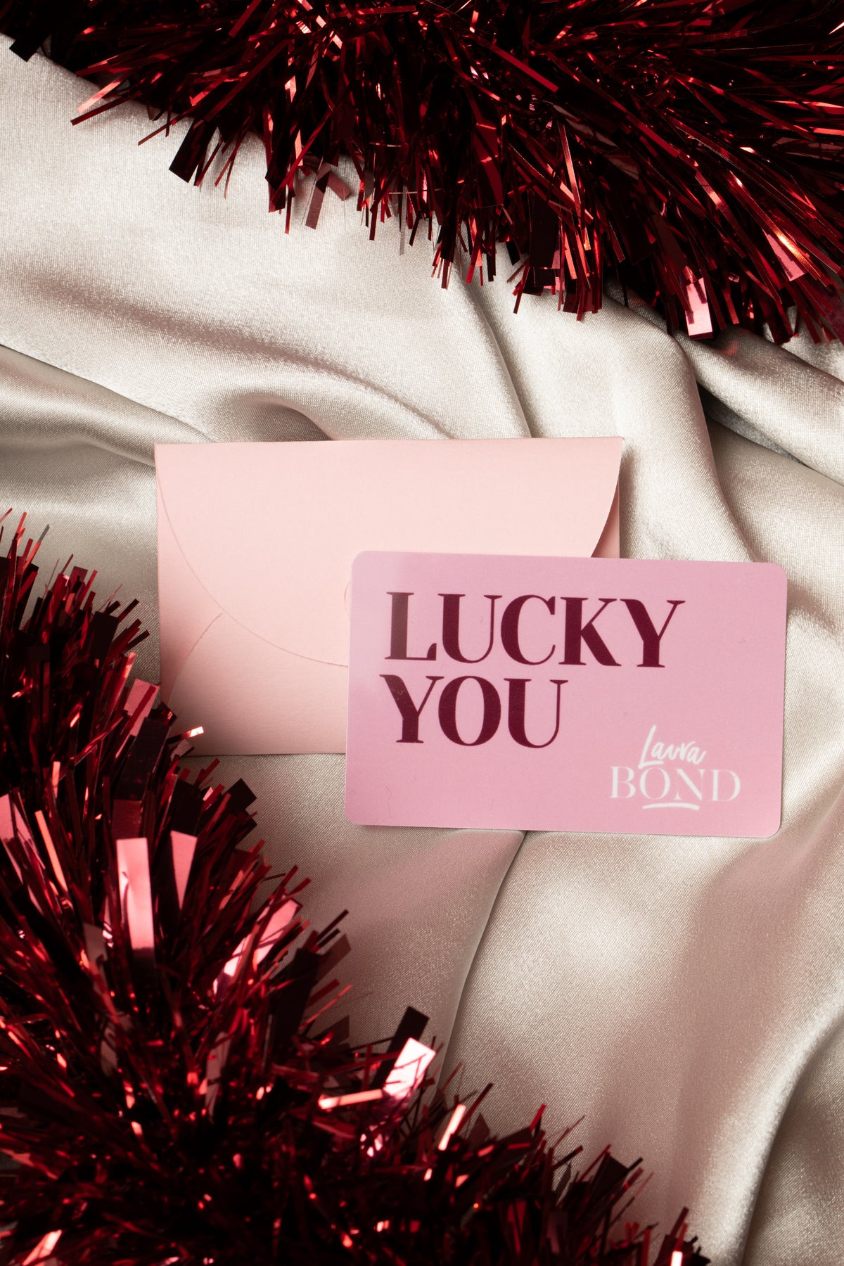 Pink Laura Bond gift card and envelope on silk with red tinsel, a Christmas gift voucher