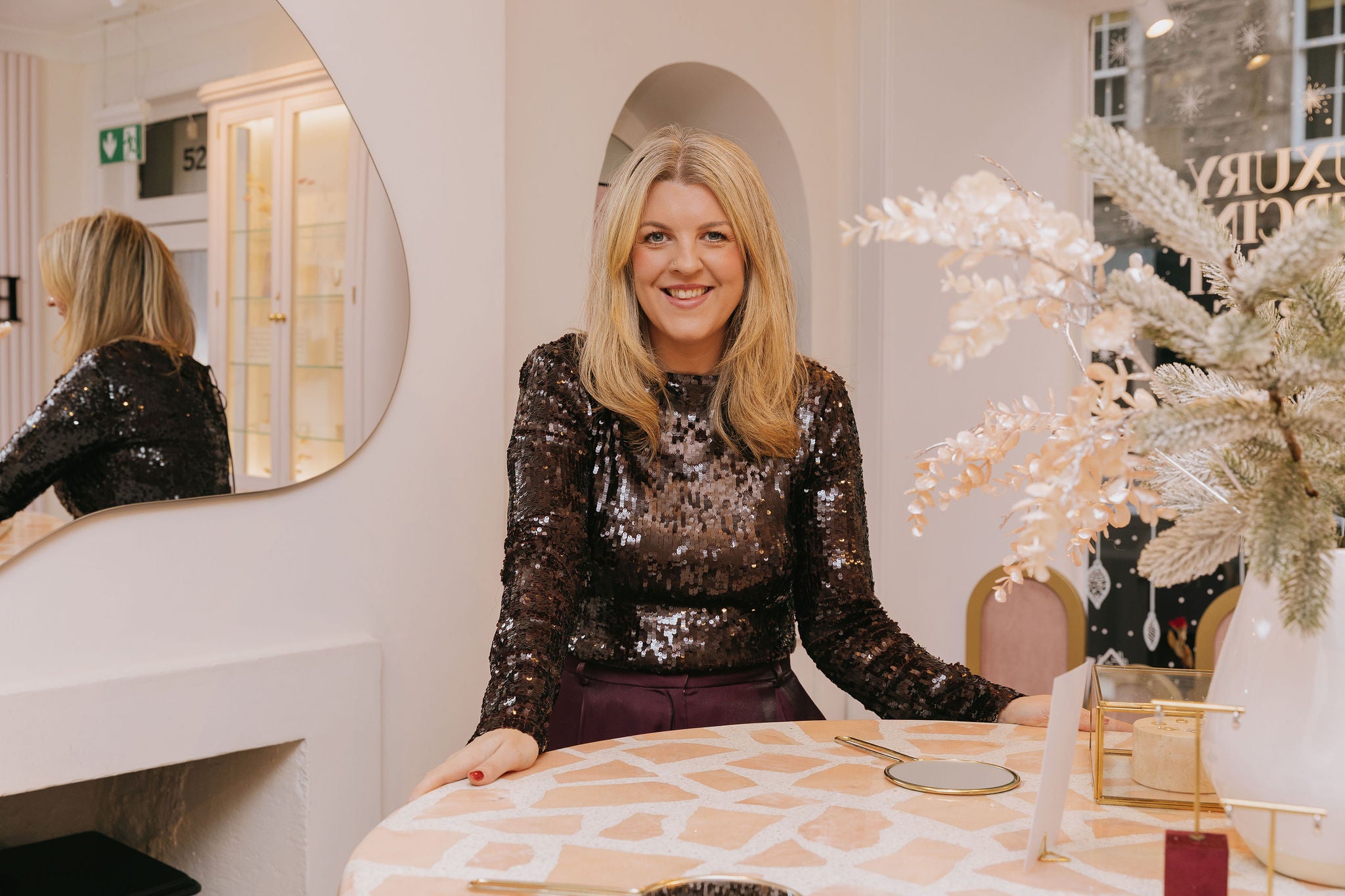 Woman in a sequin top sitting at a table with decorative items in a stylish interior setting.
