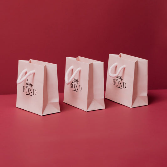 Three pale pink Laura Bond gift bags with rope handles on a burgundy background